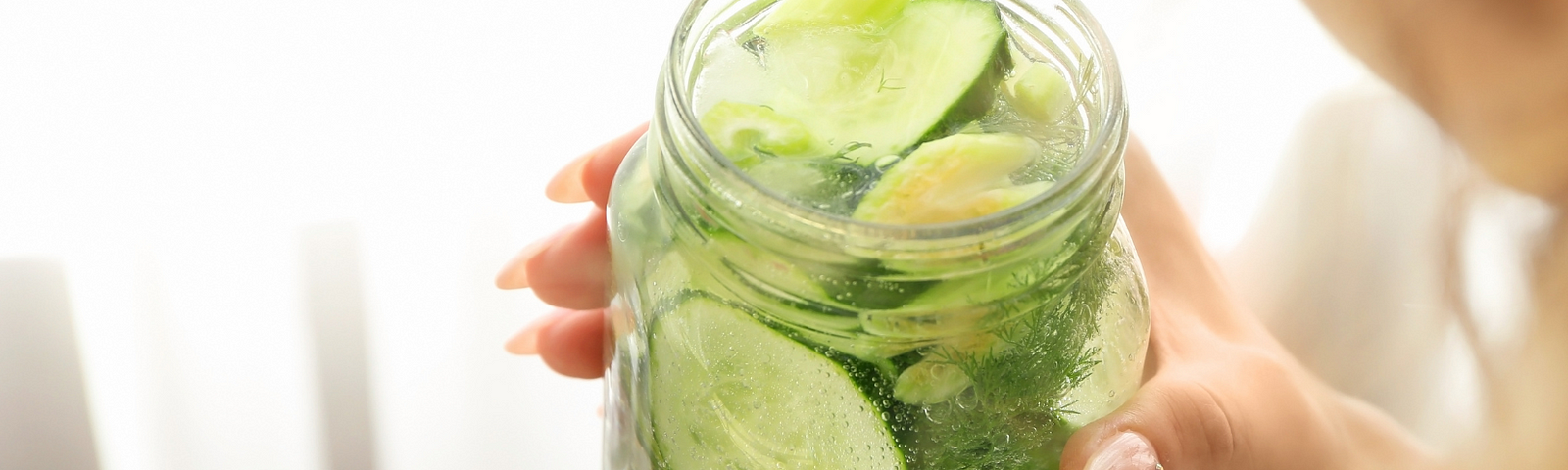 Woman’s hands holding a mason jar full of water with a whole cucumber sliced in