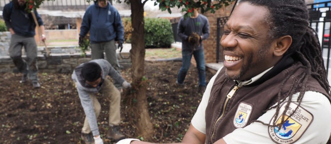 A man in a U.S. Fish and Wildlife Service uniform laughs while leaning on a shovel. Men in the background are prepping mulch around a tree.