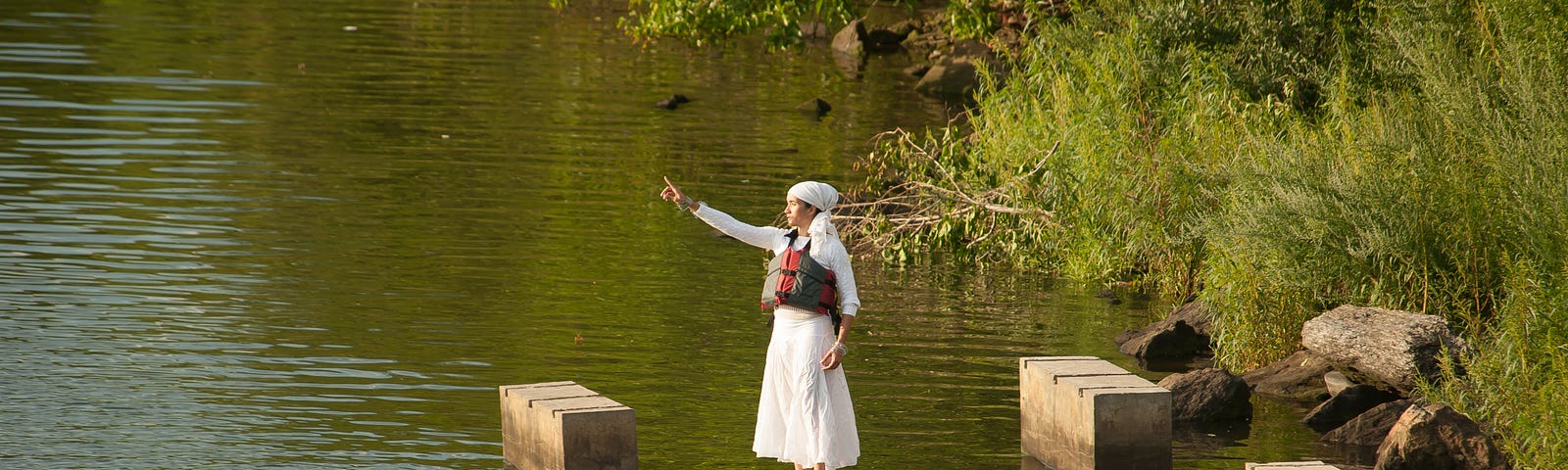 Dressed in white, woman stands on concrete pylons in Bronx River and gestures across the water, one finger pointing forward