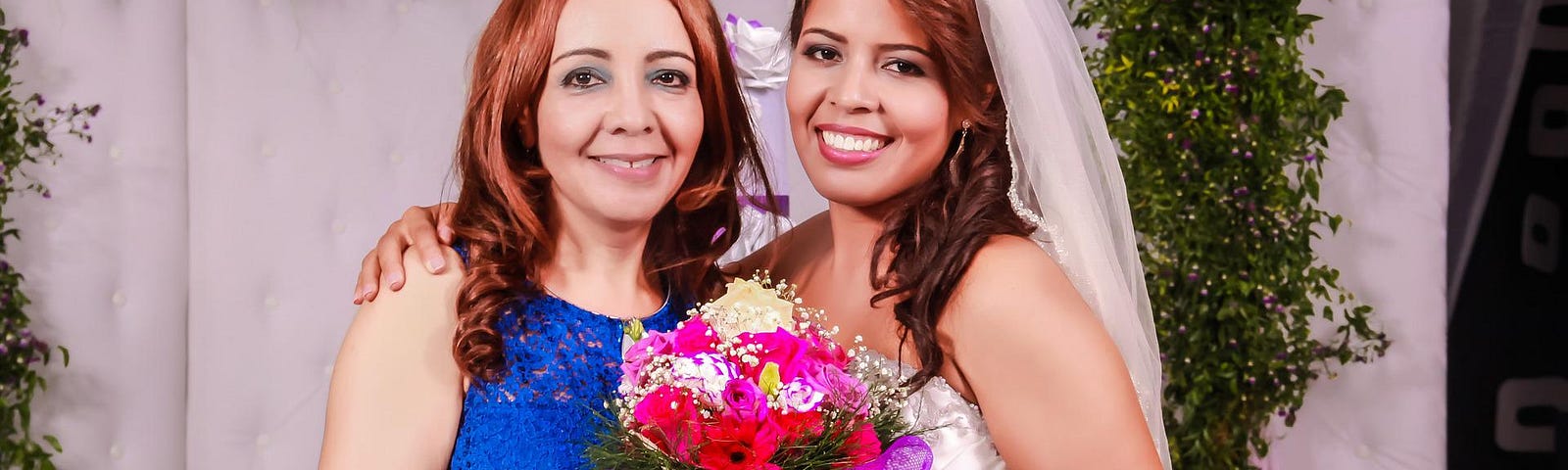 A mother of the bride poses with her wedding-dress clad daughter, who holds a bouquet of flowers. Mom wears a sapphire blue gown and a smile.