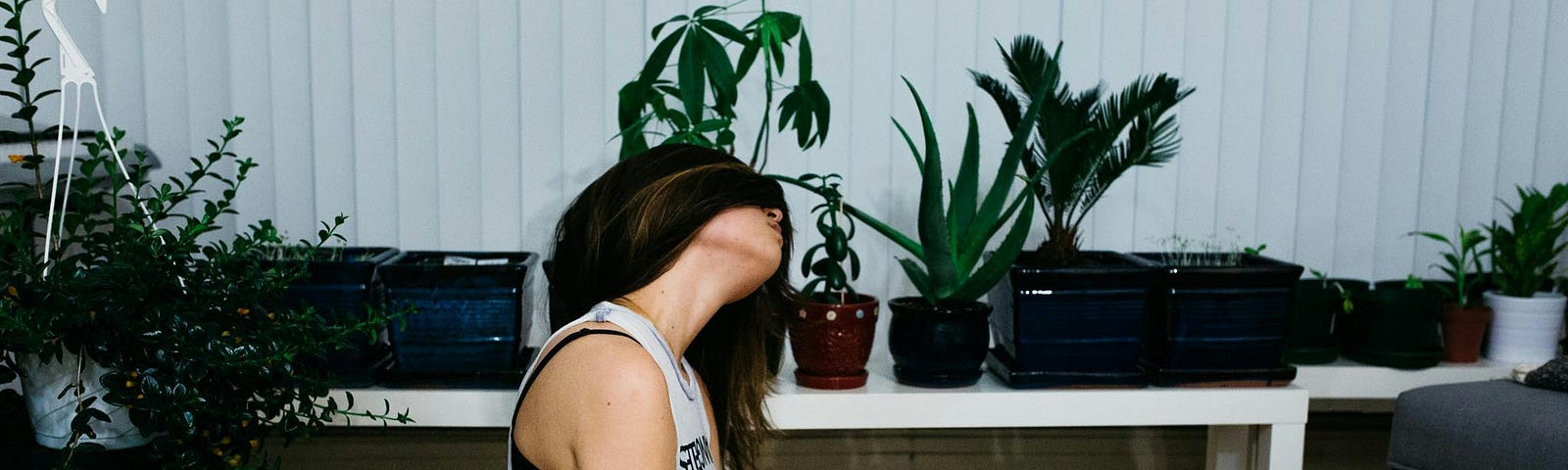 A woman sits cross-legged amid houseplants with her long brunette hair swept over her face.