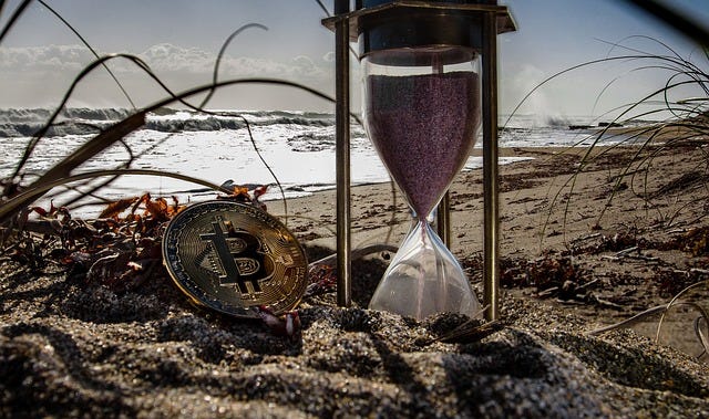 An hourglass and a bitcoin coin are on the sand on a beach. Image accompanying the Harlow Journey story, “Can the Government Shut Down Cryptocurrency?”