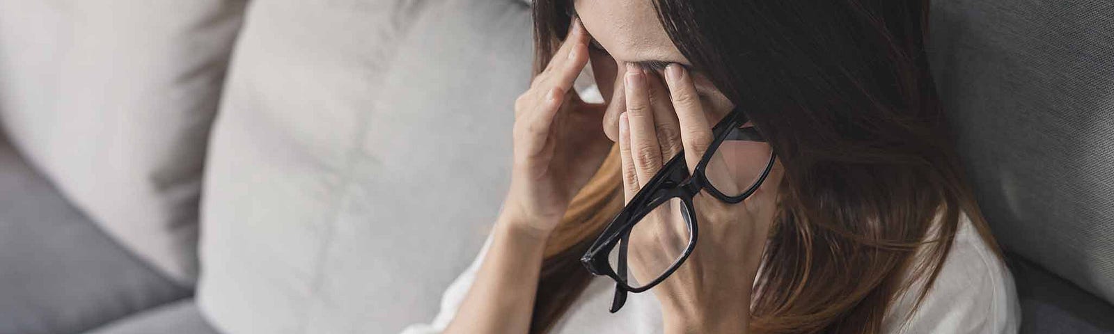 Woman holding a pair of glasses, rubbing her forehead. She’s exhausted and overwhelmed. Digital devices are nearby.