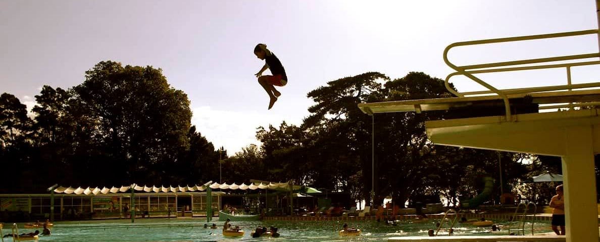 Young child jumps off diving platform into swimming pool, sun shines from behind