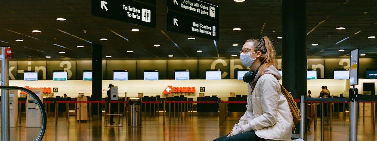 Young woman with glasses and a COVID mask, sitting on her suitcase alone, in an airport.