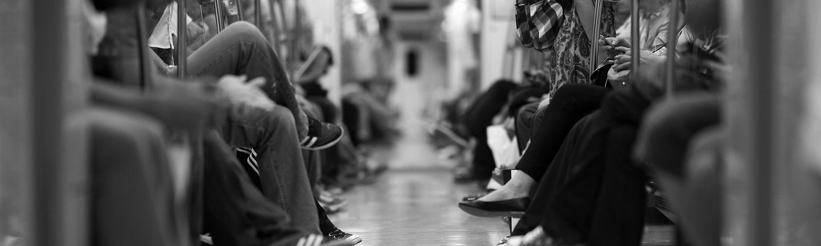 Black and white photo looking down the middle of a subway car with people’s legs on either side.