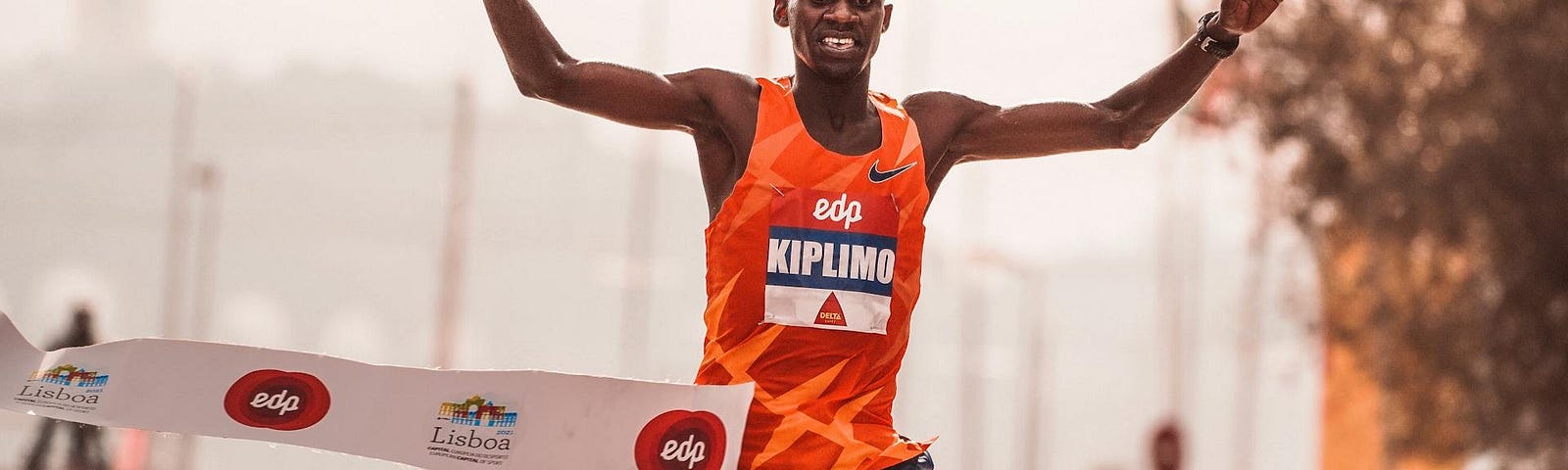 Photo of a runner finishing a race and breaking through the finish-line tape with his arms above his head and smiling proudly.