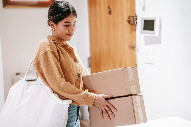 A young adult girl with a brown sweater on and a white totebag, holding two boxes in the middle of a room.