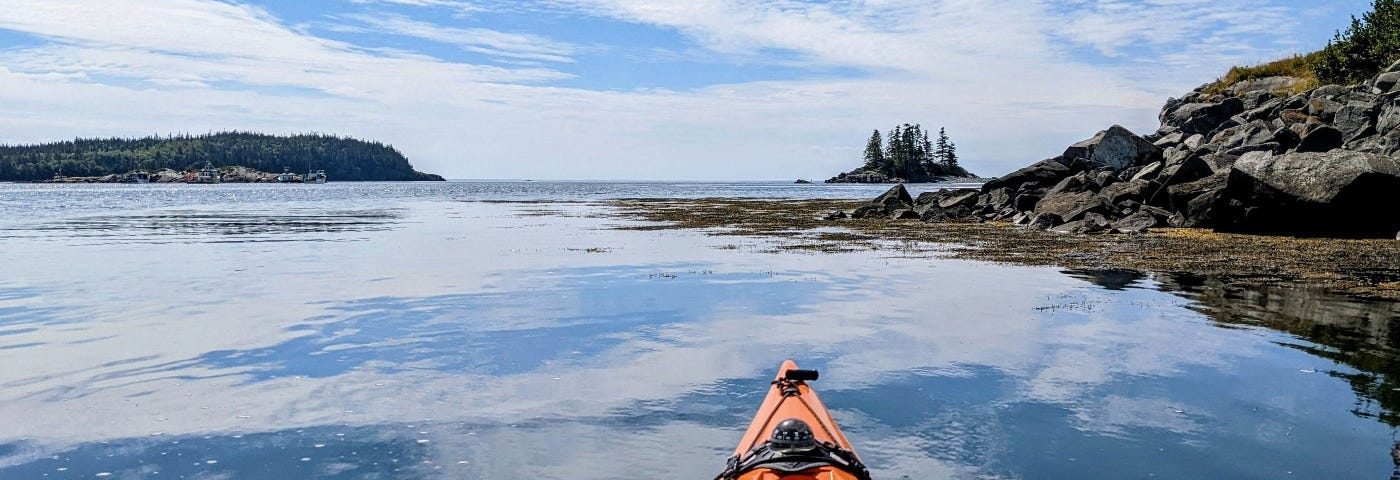 The bow of an orange kayak glides through glassy water and points toward a small island.