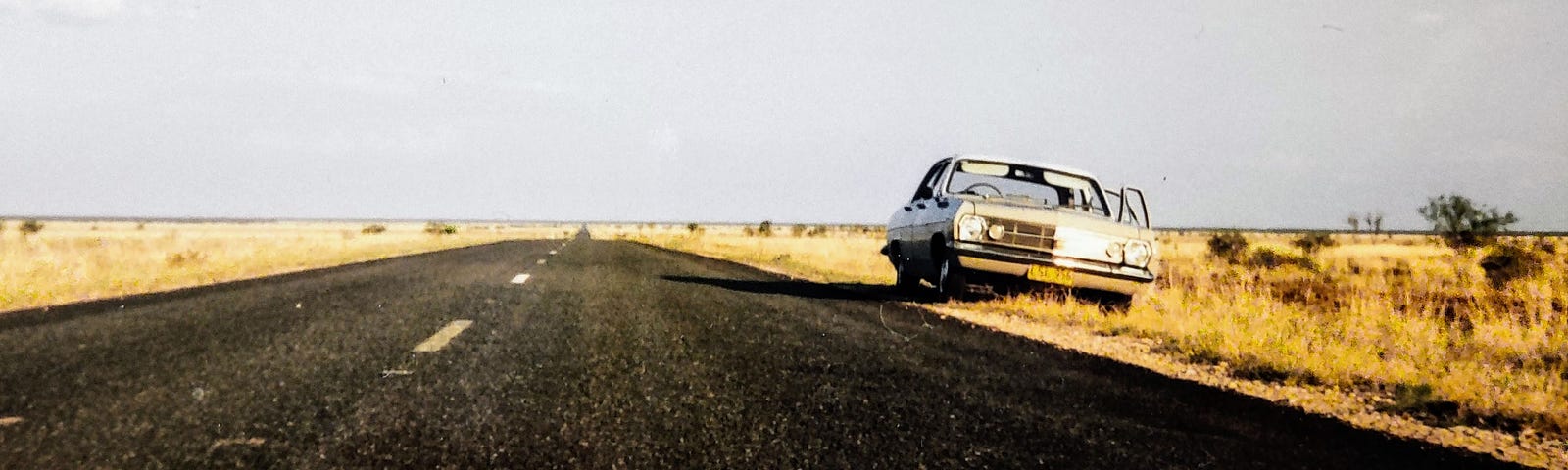 An old white car parked on the side of a desert highway in Australia, against a backdrop of blue sky and rough yellow scrubland.
