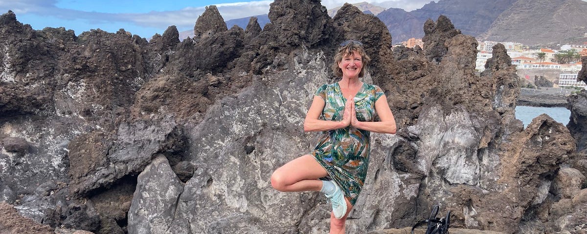I’m standing on one leg in this picture (I’m a woman in my fifties). There are volcanic rocks behind — this is Tenerife — and cliffs in the distance, and a glimpse of blue sea.