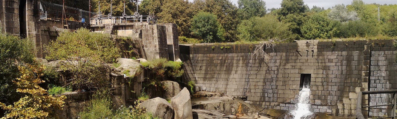 View of the Amieiro Galego dam in Vila das Aves Portugal. A dead willow tree can be seen hanging on top of the stone wall over the dark waters river stream.