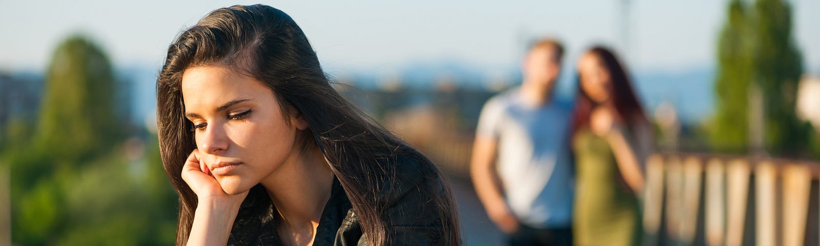 A young woman standing on a bridge, leaning on her hand and looking upset. Behind her, an out-of-focus couple stands arm in arm, clearly having a good time together.