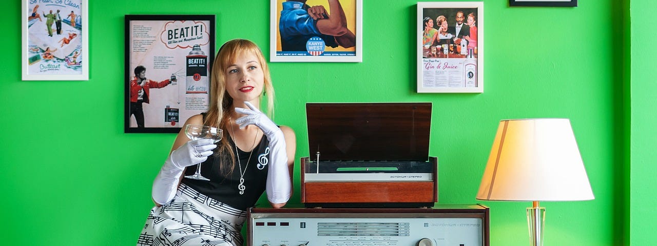 A 1960’s blonde girl sitting next to a turntable and stereo holding a drink in white gloved hands