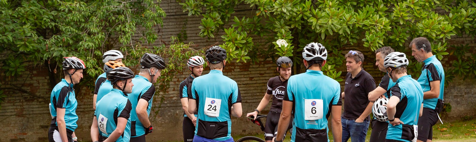A dozen people wearing blue cycling tops and helmets stand in a circle as they listen to a man holding up a bike.