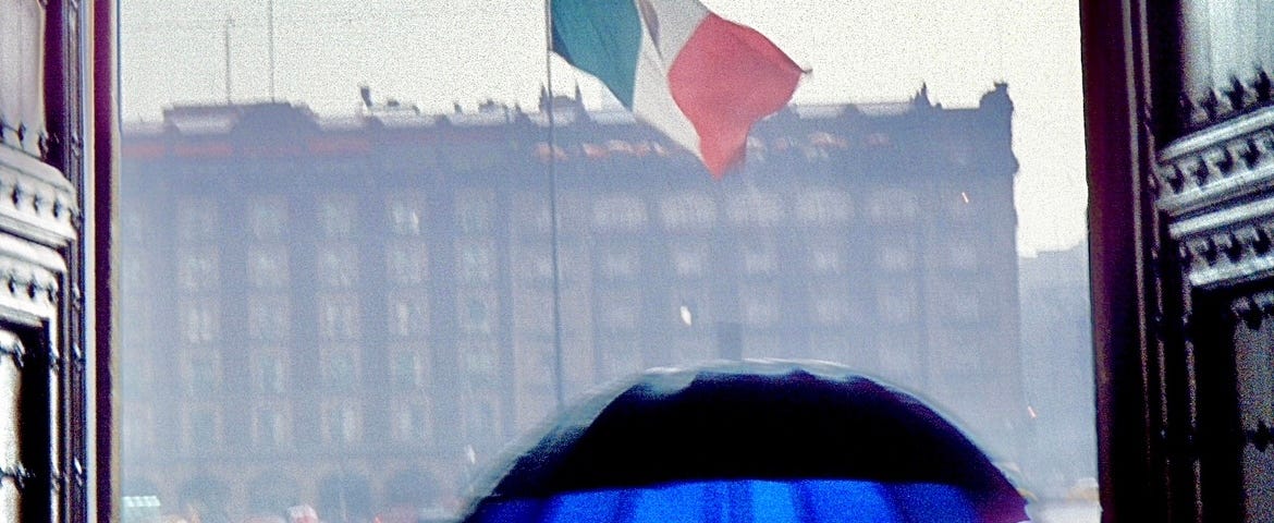 A Mexican flag blows in the wind in a large open square during a rainstorm, photo is taken through large wood antique doors of the National Palace.