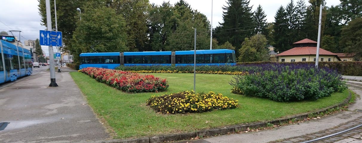 A blue tram is parked next to an empty sidewalk by a spacious green park entrance