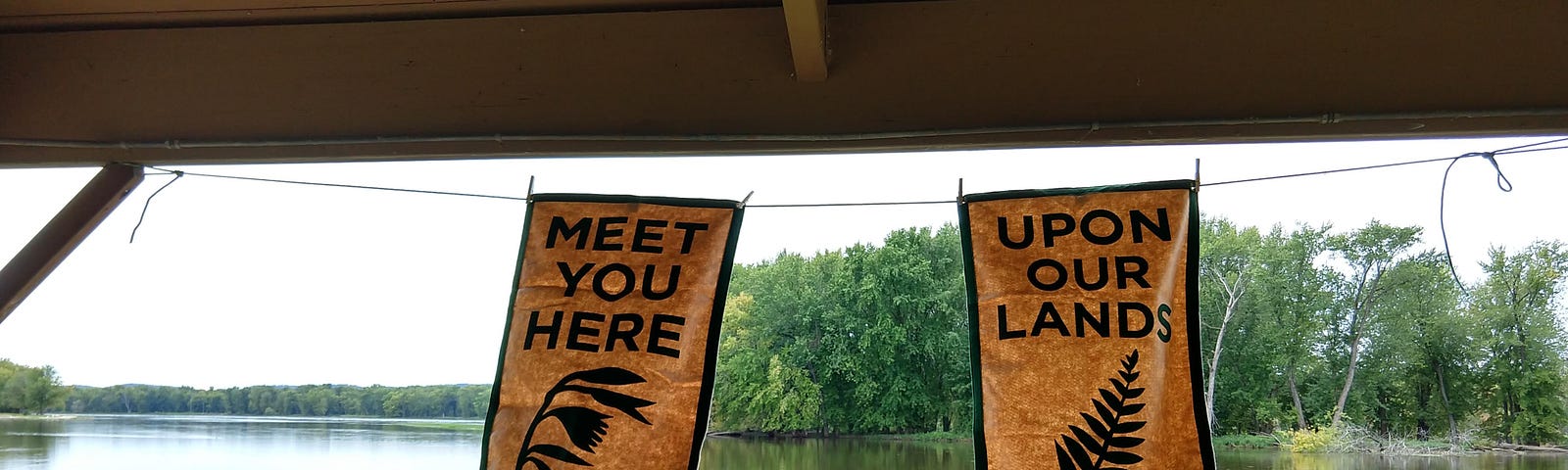 Two banners reading “Meet you here” and “Upon our lands” in picnic shelter near river.