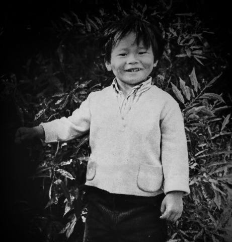 A monochrome portrait shot of a smiling, shy, five-year-old boy standing next to some tall bushes.