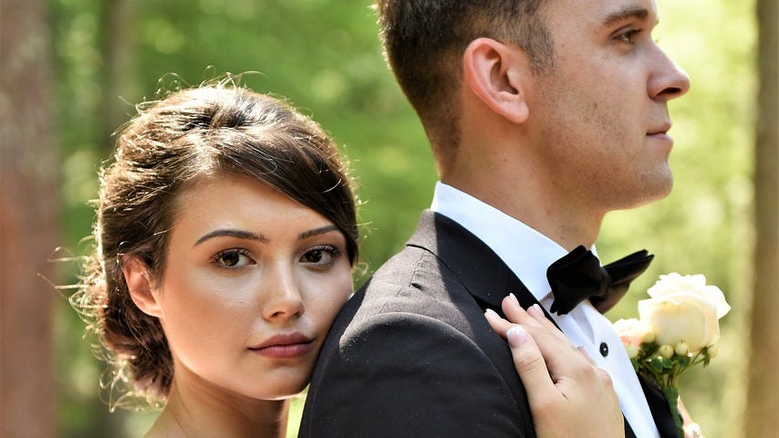 Image of a couple on their wedding day — the bride is a brunette in a white dress and she stands behind the groom, resting her cheek on his shoulder and her hand on his chest. He has short hair and wears a dark suit and bow tie with a white rose in his buttonhole