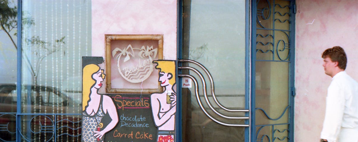 A colorful cartoon female & male in their swimming suit illustration on the menu board outside a seaside restaurant. The chef in his white uniform just arrived on the right-hand side of the photo, looking at the cartoon figures.