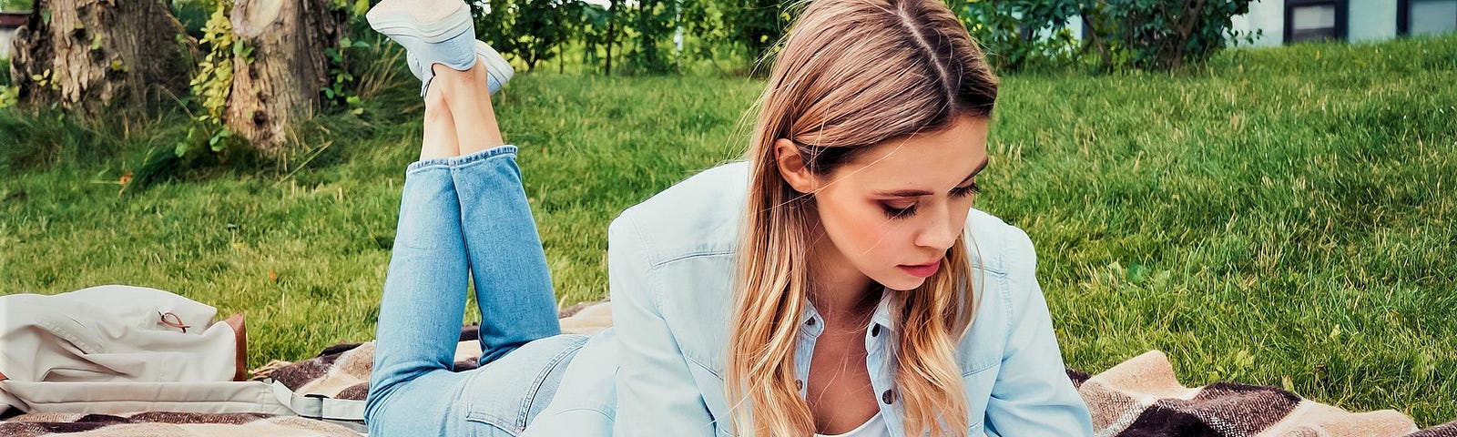 with with long blonde hair laying on blanket on grassy lawn reading a book