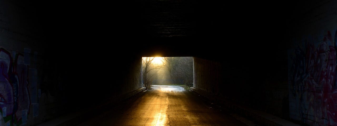 The viewer seems to be inside a dark tunnel. Light shines in the other end of the tunnel, creating a golden path forward.