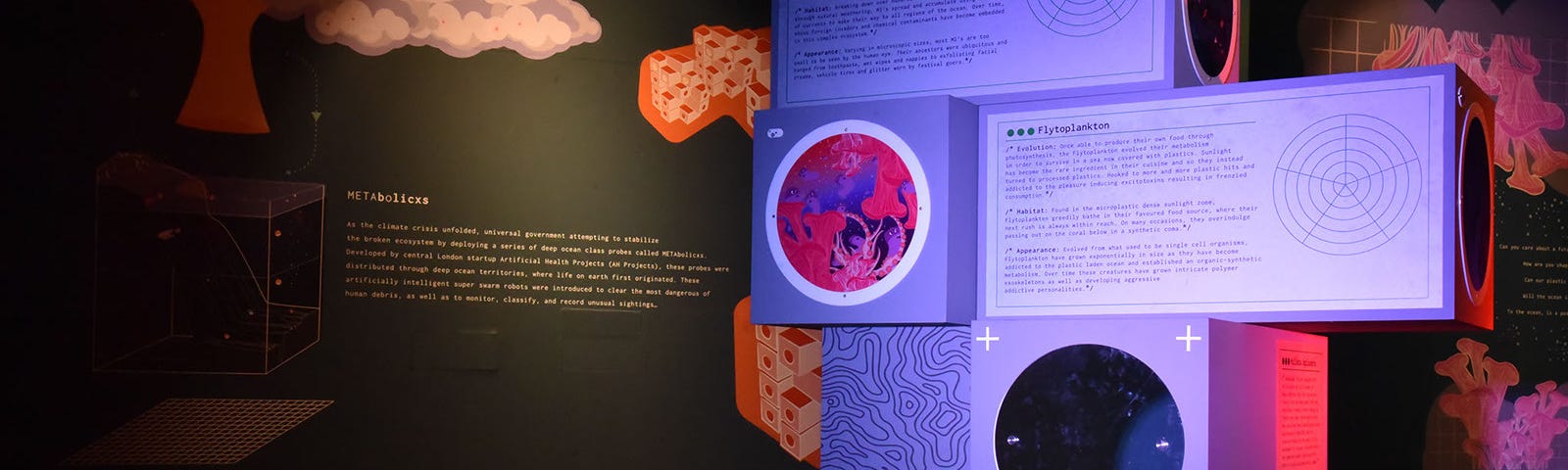 This is a photo of an art installation titled The Anxious Ocean and the Moonbathers by Hyphen Labs. Well lit purple rectangular boxes in the foreground with circular drawings and typography on them amidst what appears to be fabric on the floor. There are illustrations painted on darker walls.