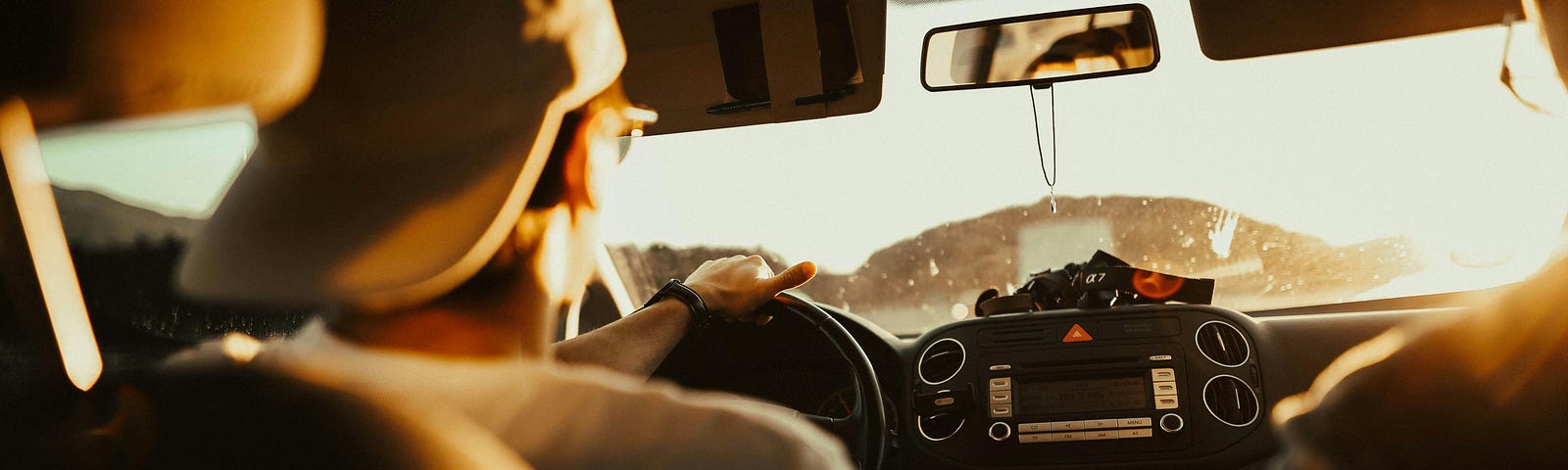 Photo of a male-presenting driver wearing a baseball cap and glasses sitting at the steering wheel of a car, taken from behind