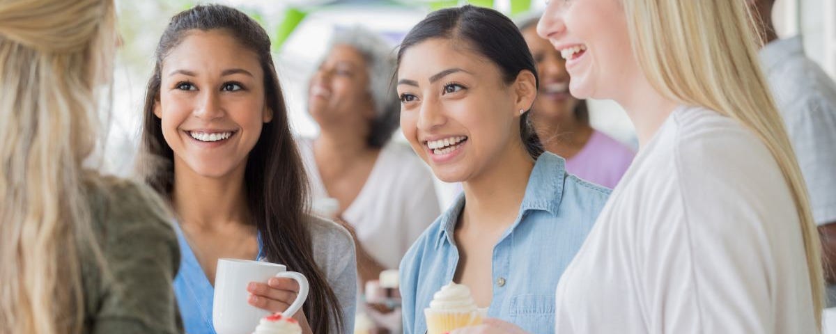 4 women standing talking and smiling each holding a white coffee mug and white plate with a muffin.