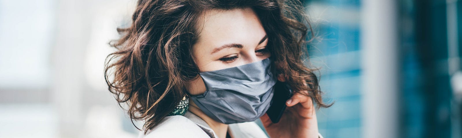 A stylish woman with short brown hair smiles behind a mask in the city.