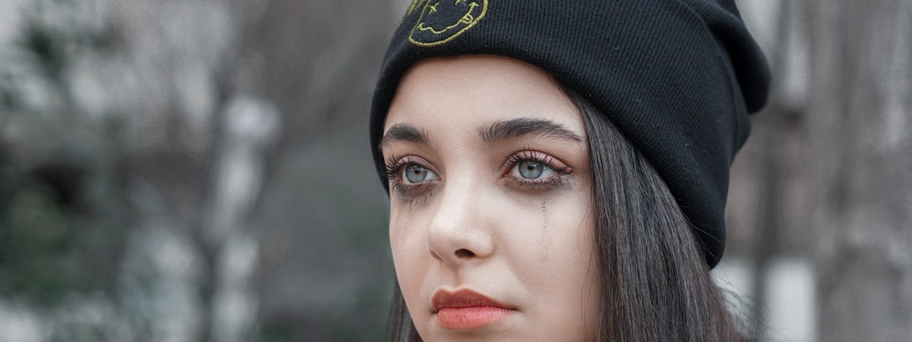 Young woman wearing with tearful eyes wearing a shirt and black cap against a blurry outdoor background