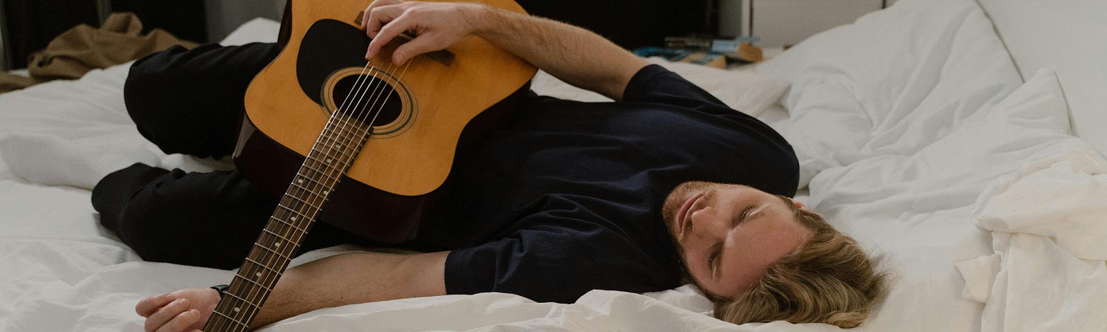 Image shows man with blond hair and dark clothing lying despondently on a bed with white linens while holding and strumming an acoustic guitar