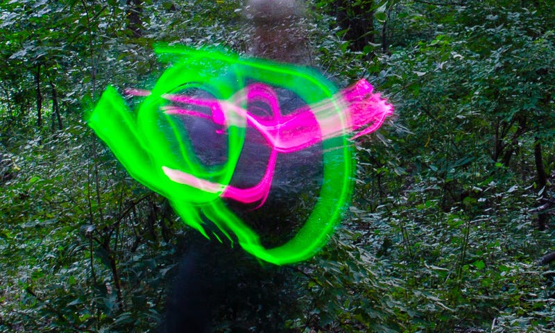 A dark forest background, a blurry person figure with green and red light streaks about the head and shoulders area. Apparently standing on one foot with the other foot being held up in the air. There’s a yellow circle around the ankle of that foot. A firefly.