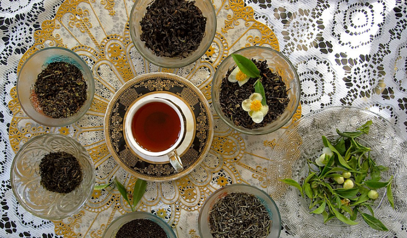 A photograph of six glass containers with loose tea, one with flowers and leaves. There is a cup of tea and saucer in the middle. To the side is a plate of leaves and light green buds. The teacup, containers of tea, leaves and buds are placed on top of a white lace cloth and a gold lace doily.