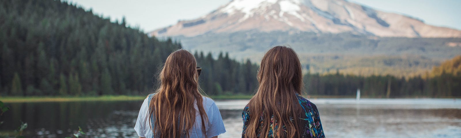 Two young women are sitting on a ledge of a stone wall, overlooking a lake. Their backs are faced toward the camera. Both are wearing comfortable clothing and have long brown hair that falls down their back. The view in front of them is an ascending forest of lush pine trees and a great snow-capped mountain in the distance.
