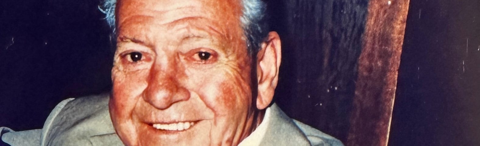 A close-up of an old man smiling at the camera. His face is red and blotchy, his eyes bloodshot, but his false teeth are white. His grey hair is sleeked back with hair gel. He wears a fawn suit that looks too big across his narrow shoulders, with a white shirt and black tie.