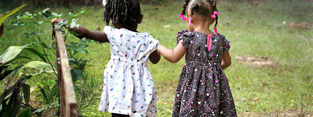 Two little girls, with patterned dresses and curly hair, hold hands as they venture into a garden.