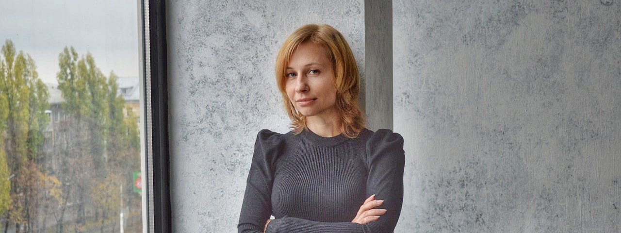 A confident woman in a grey dress stands by a wall.