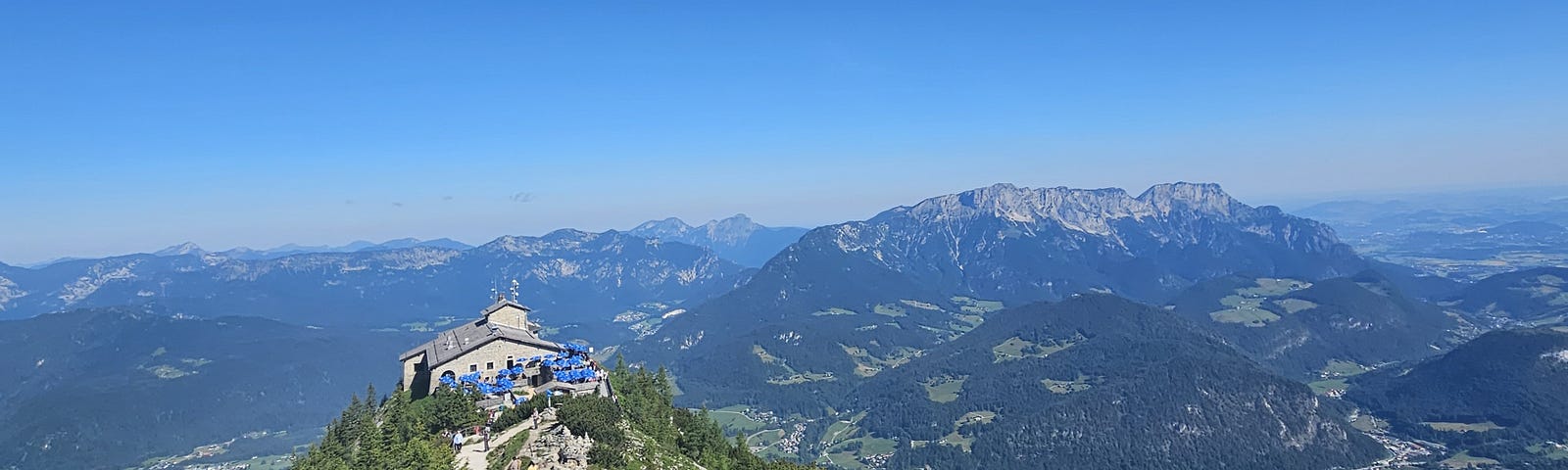 A chalet sits on a mountaintop, a path leading up the mountain from it. Alps rise in the background