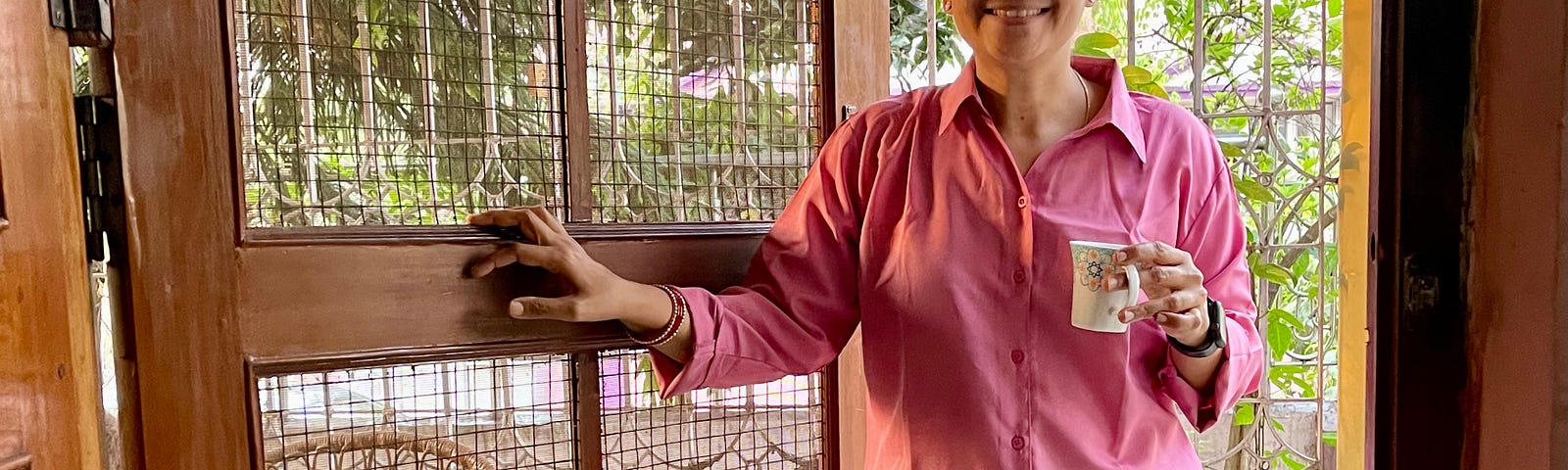 A woman wearing a pink, full-sleeved shirt with a collar and black trousers is holding a coffee cup and is smiling at the camera.