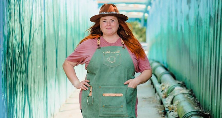 A woman with red long hair wearing a hat, green apron, and a smile.