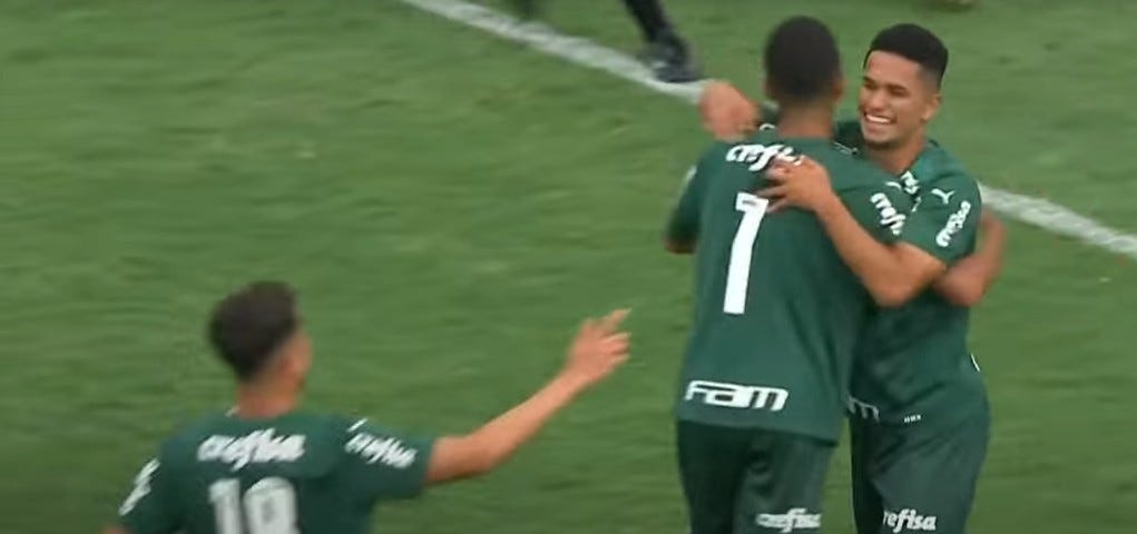 Jogadores do Palmeiras festejam gol contra o Assu pela Copa São Paulo de Futebol Júnior. À direita, Gabriel Silva, de frente, abraça Giovani, que está de costas e usa a camisa número 7. Outro jogador, com o número 18, chega para abraçar e aparece com a camisa 18. Todos estão cortados da cintura para cima. Usam uniforme verde do Palmeiras.