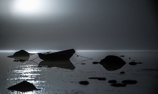 Moody black and white photo. A smooshed blob in the sky is the moon. In dark silhouette on the surface of water is a small boat tied to several large rocks.