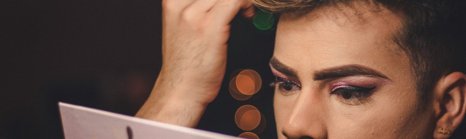 A performer applies makeup while using the mirror on their blush pad. Their eyebrows are slightly furrowed. They appear deep in concentration. They have colorful eyeshadow and winged eyeliner.