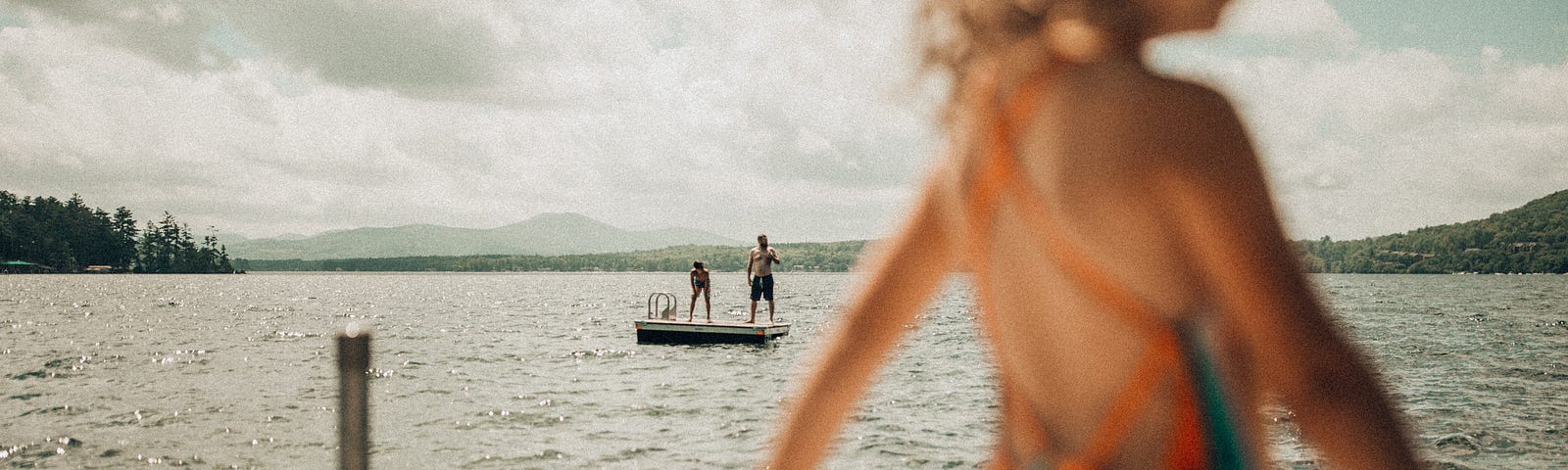 Child in a striped bathing suit standing by a lake in the foreground with two older people standing on a floating raft in the background.