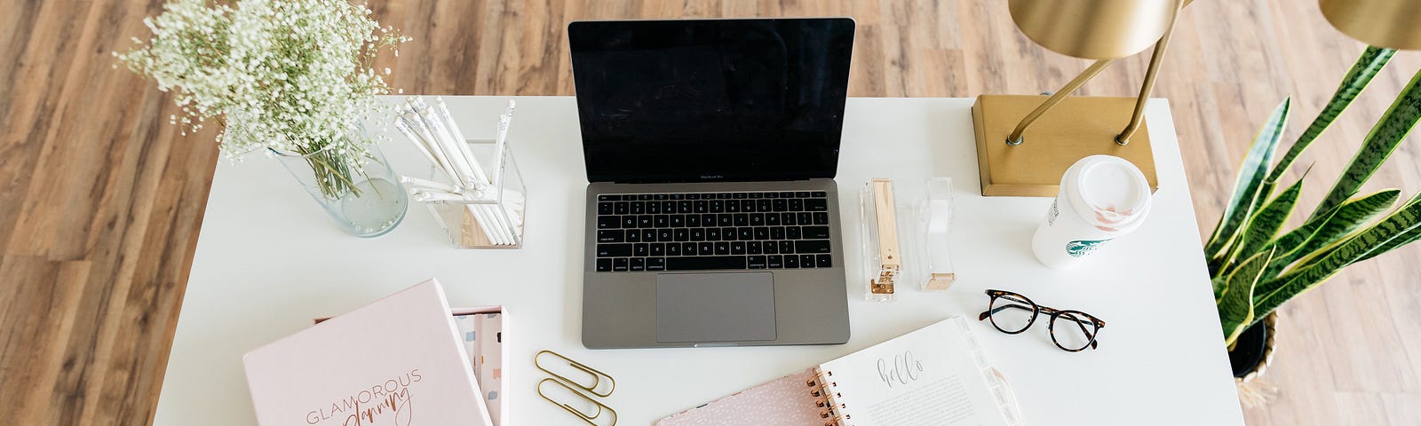 A laptop, diaries, paperclips , glasses a plant and some flowers on a desk