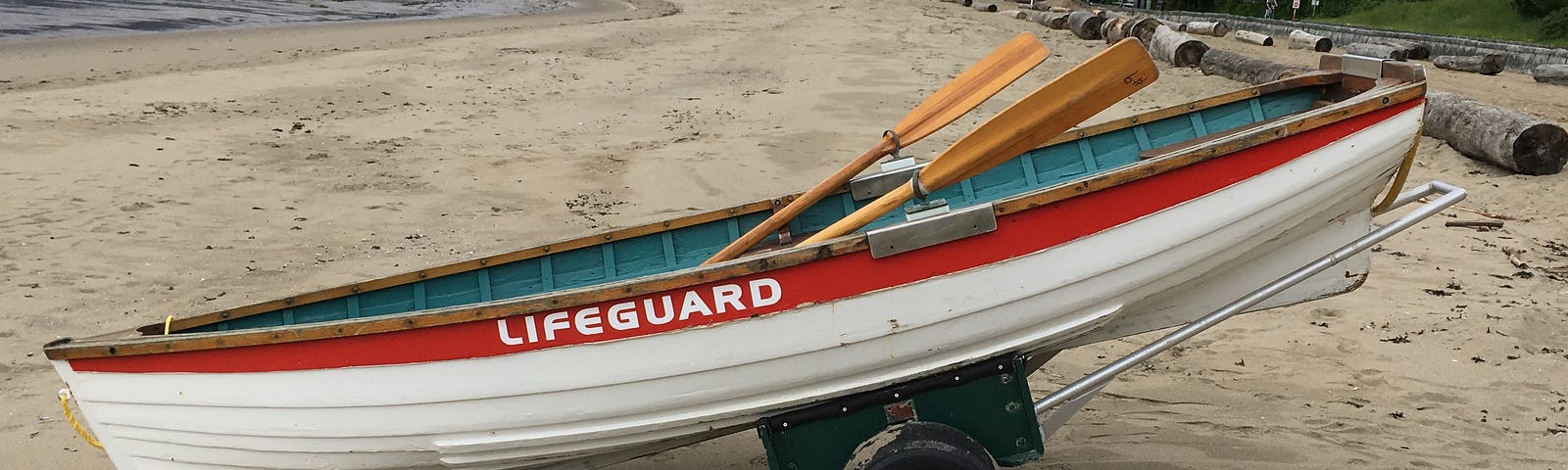 A photo of a small white dinghy with a red line painted on the top wooden panel and the word Lifeguard in white letters. There are two oars in the boat and it sits on a sandy beach.