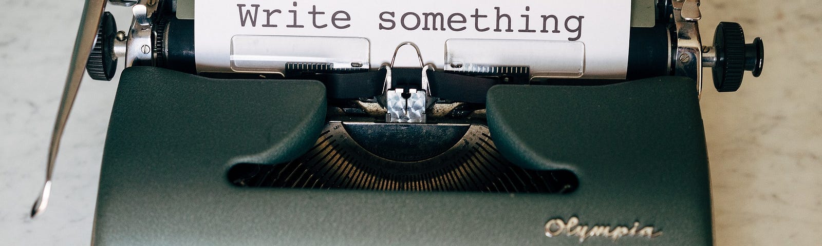 Old school typewriter on a desk with a piece of paper rolled into it. The paper reads: “write something”