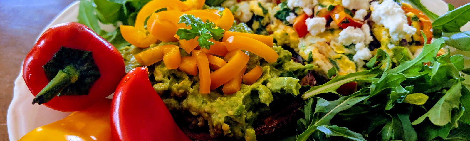 White plate of brunch food including scrambled eggs, avocado toast, fresh arugula and mini sweet peppers .
